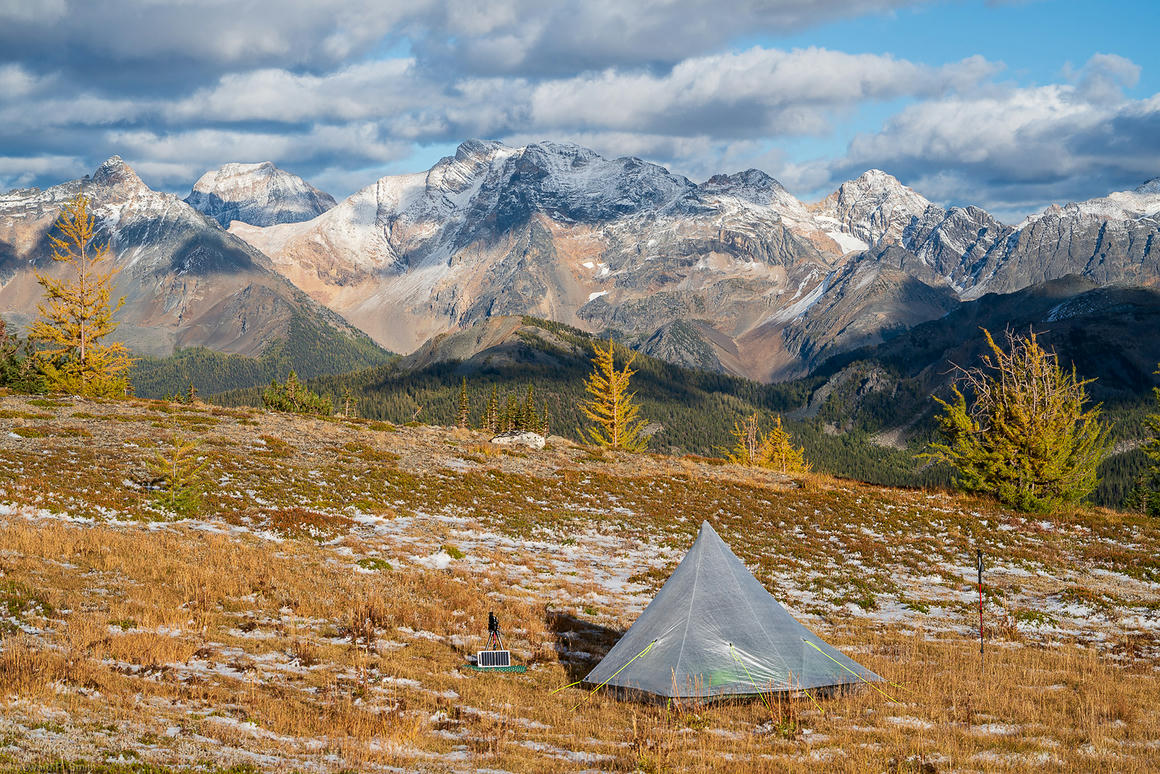 Alpine camping setup with a small tent and solar panel on a snow-dusted ground, with the Septet mountains in the background under a cloudy sky.