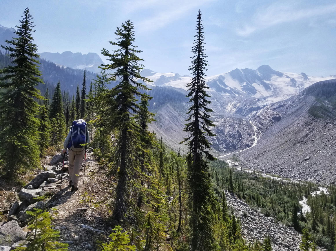Backpacker ascending a glacial moraine in the Purcell Mountains during a photography trip, with towering conifers framing the rugged landscape