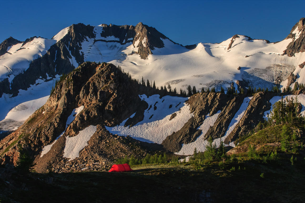 A solitary red tent pitched in an idyllic alpine setting near the Bugaboos, with a stunning backdrop of snow-covered peaks illuminated by the summer sun, perfect for a photography trip