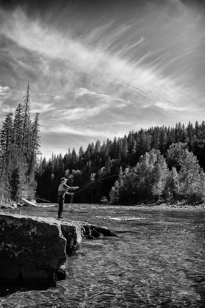 Jim fishing on the riverbank, surrounded by lush forest under a sweeping sky, in a tranquil moment that balances the demands of work