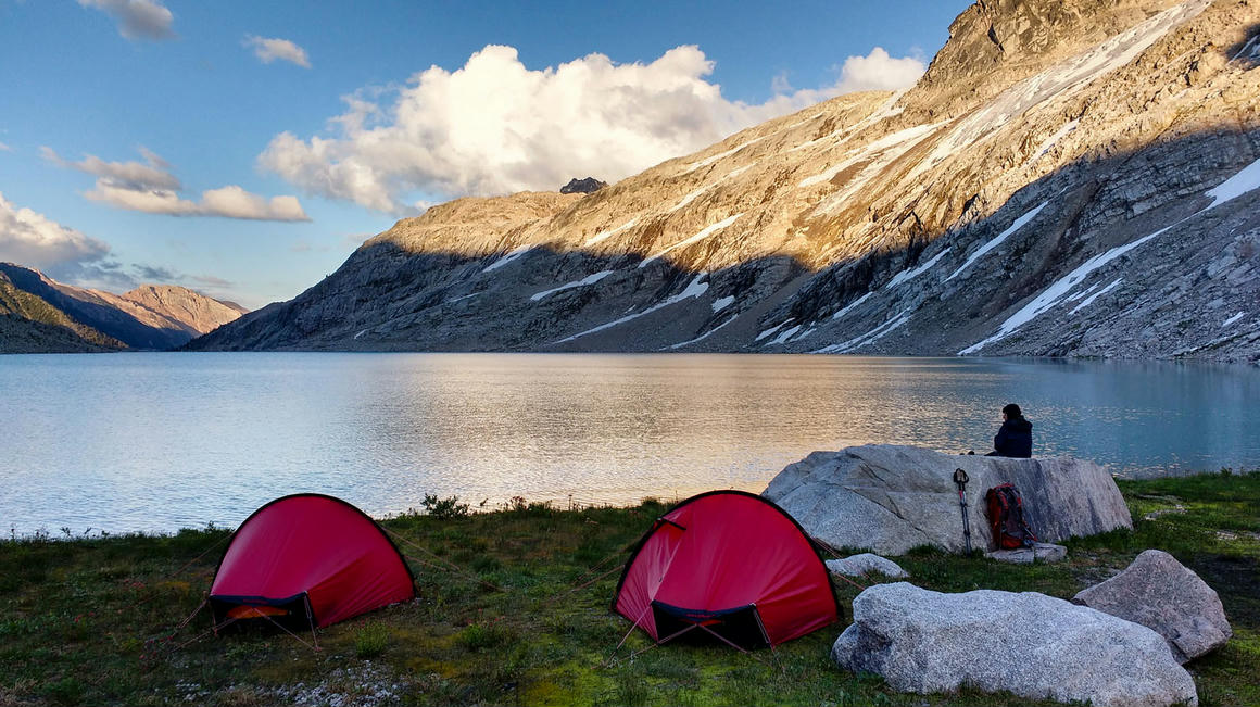 Camping by a glacial lake in the Canadian Rockies with two red tents and Jim Smith seated on a boulder, contemplating the serene waters at sunset
