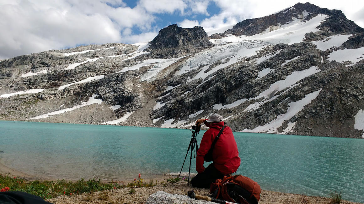 Howard Smith, photographer, setting up a tripod to capture a high-resolution panorama of a turquoise glacial lake and rugged mountain terrain in the Canadian Rockies