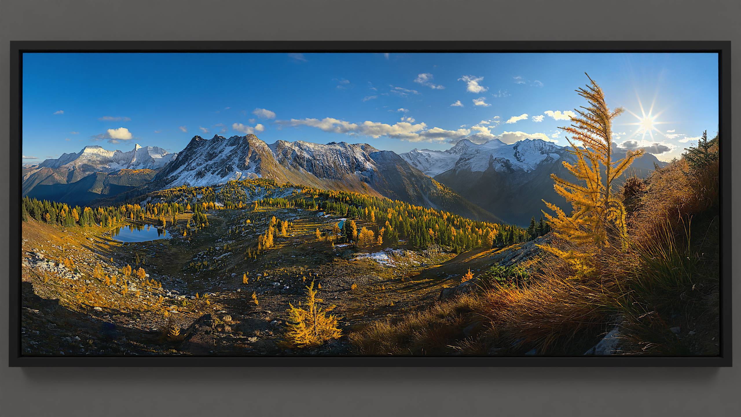 An autumn evening at Jumbo Pass reveals golden larch and fir trees glowing against a backdrop of Bastille mountain's light-dappled face. Below, a pond reflects the sky while Jumbo cabin nestles atop the pass, overseen by a vibrant sun.