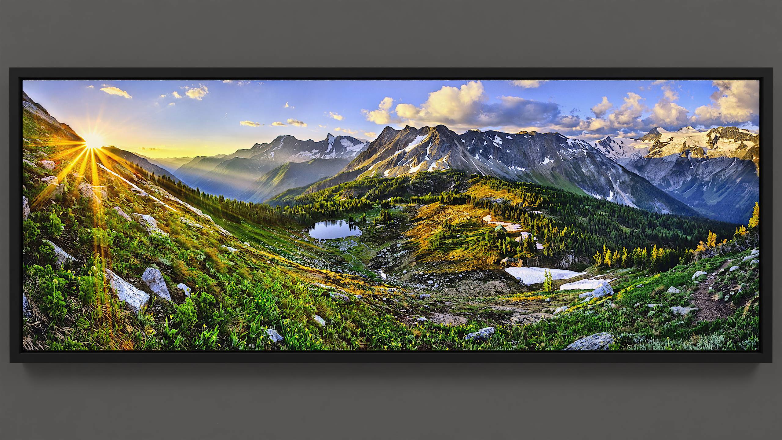 A panoramic scene unveils a spring morning on Jumbo Pass, atop an alpine hillside carpeted with fluttering glacier lilies. The rising sun, a radiant star, casts warm rays across distant blooms and the valley. The mountain pass below bathes in early morning light, where trees craft patterns of shadow and illumination, beside a placid lake reflecting the azure sky. A solitary cabin basks in the sunlight. Beyond, Bastille mountain glows gold, with the distant Horseshoe glacier sparkling under the sunlight. Overhead, a brilliant blue sky hosts gently lit, billowing clouds. A spectacle that rewards the early riser.
