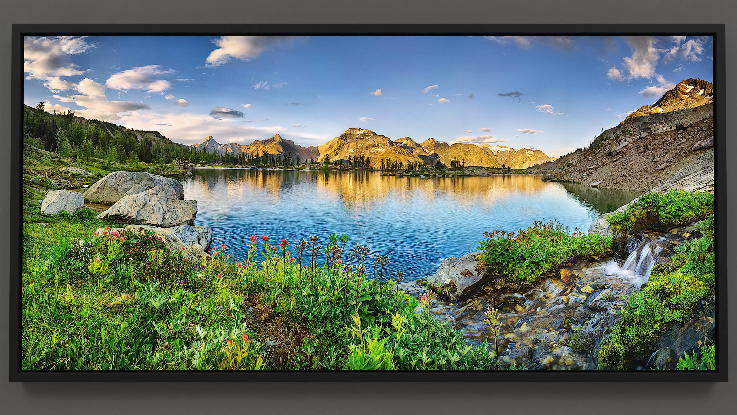 Evening settles over an alpine lake, where a splashing stream cascades past blooming red Paintbrush wildflowers. Granite boulders line the lakeshore, reflecting alongside distant silhouettes of larch trees and the Septet Mountains under a sky of silky clouds.